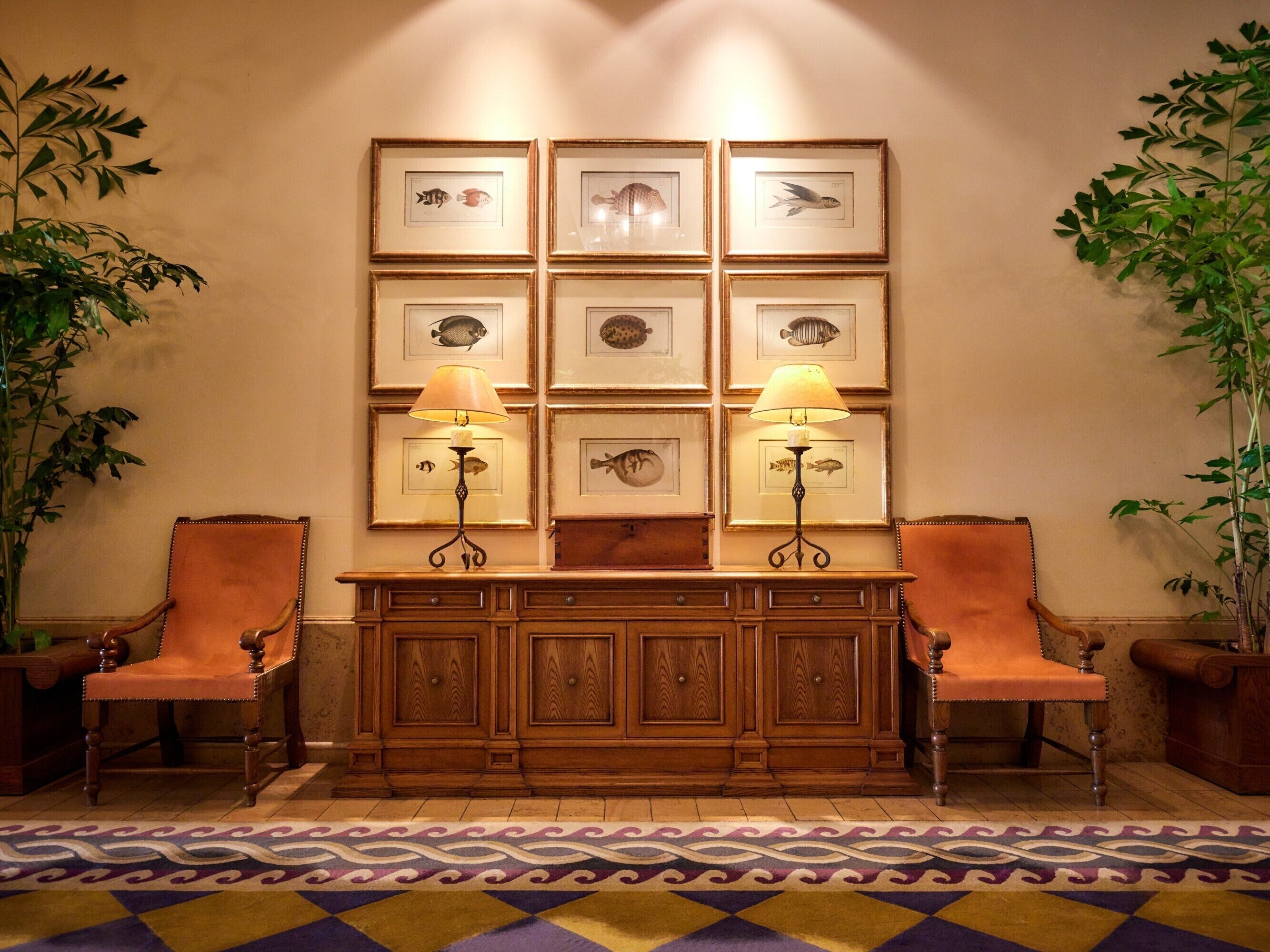 A lobby with a wooden console table flanked by two leather chairs, a wall adorned with framed fish illustrations, and two floor lamps.