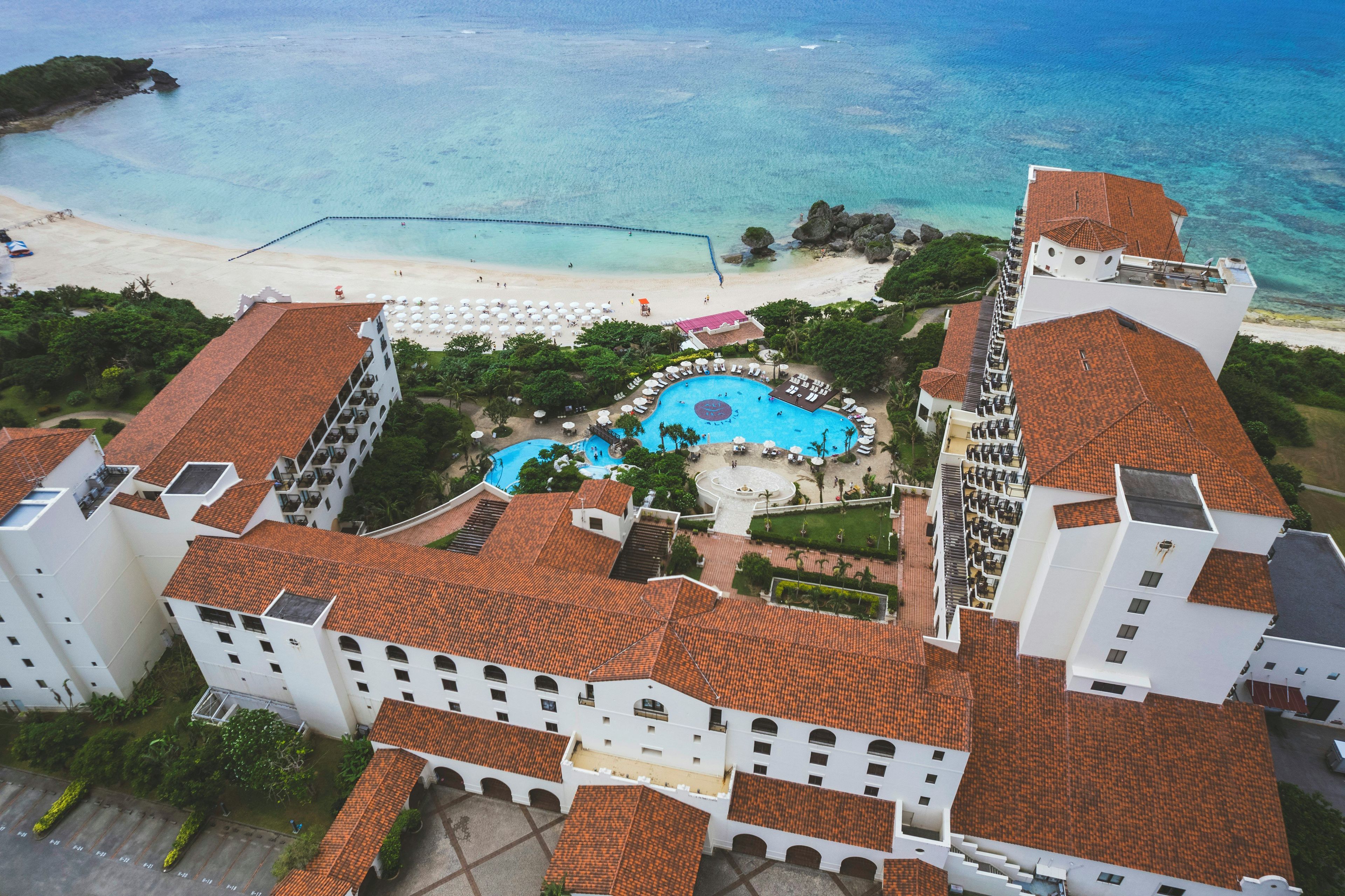 Un resort con una piscina grande, acceso a la playa y varias edificaciones con techos de tejas rojas.