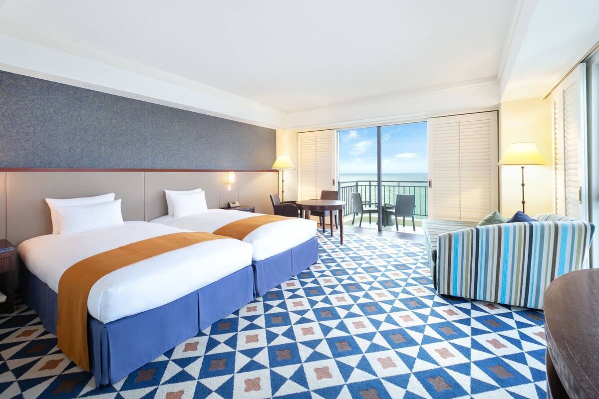 A hotel room with two beds, a balcony with a view, a dining area, and a patterned floor.
