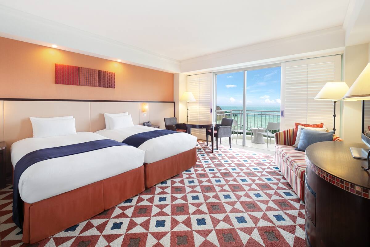 A hotel room with two beds, a balcony with a view, a desk, and a sofa.