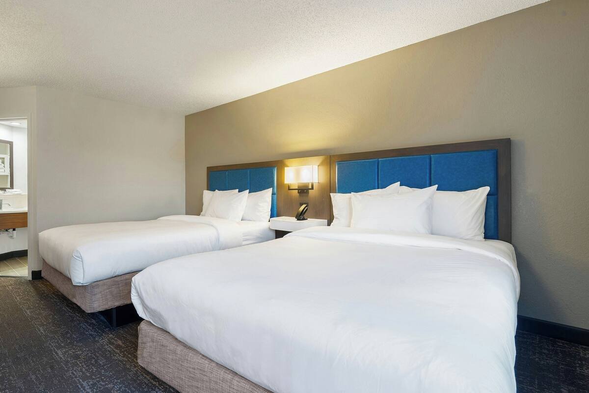 A hotel room with two beds, a nightstand with a lamp, and a bathroom visible in the background.