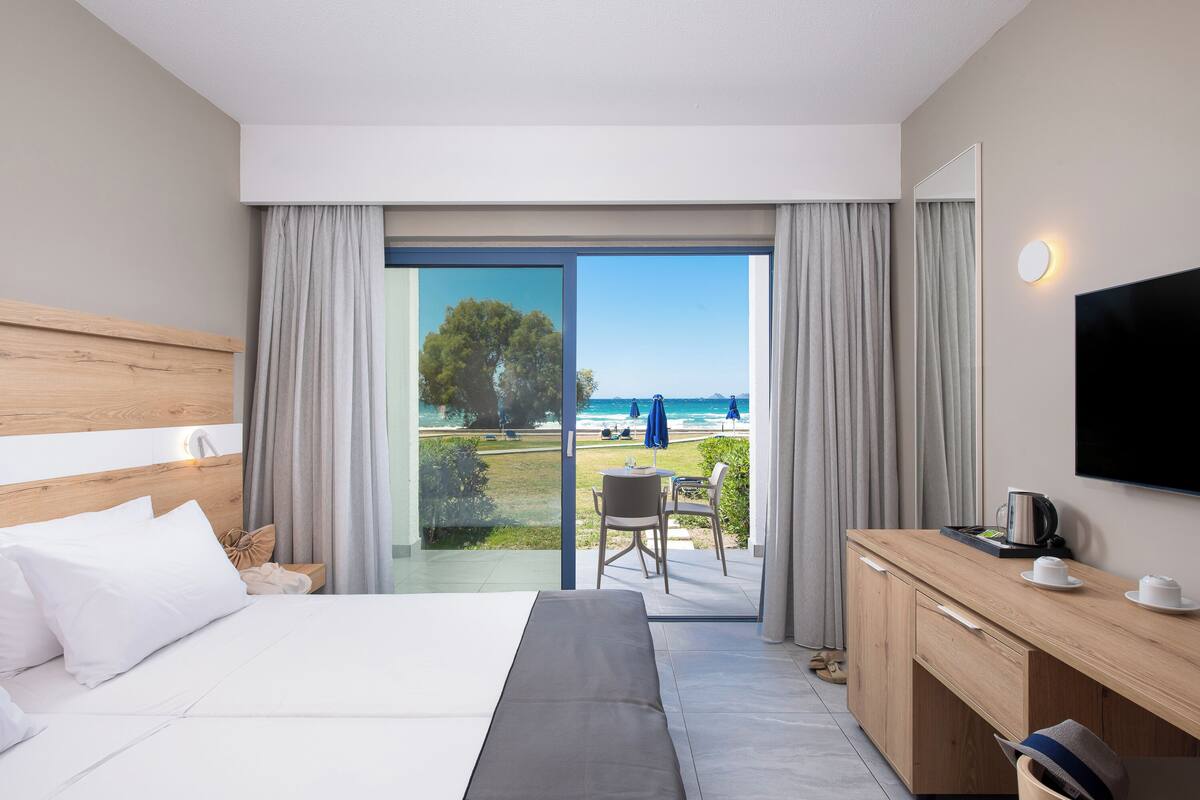 A hotel room with a bed, a desk, a TV, and a view of the beach.