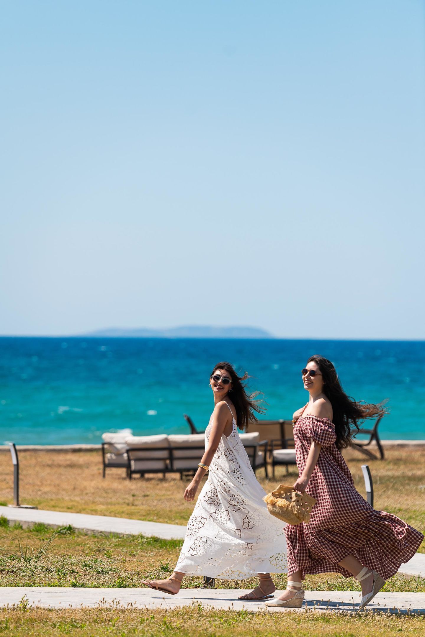 Two women walking on a path by the sea with a clear view of the ocean and distant land.