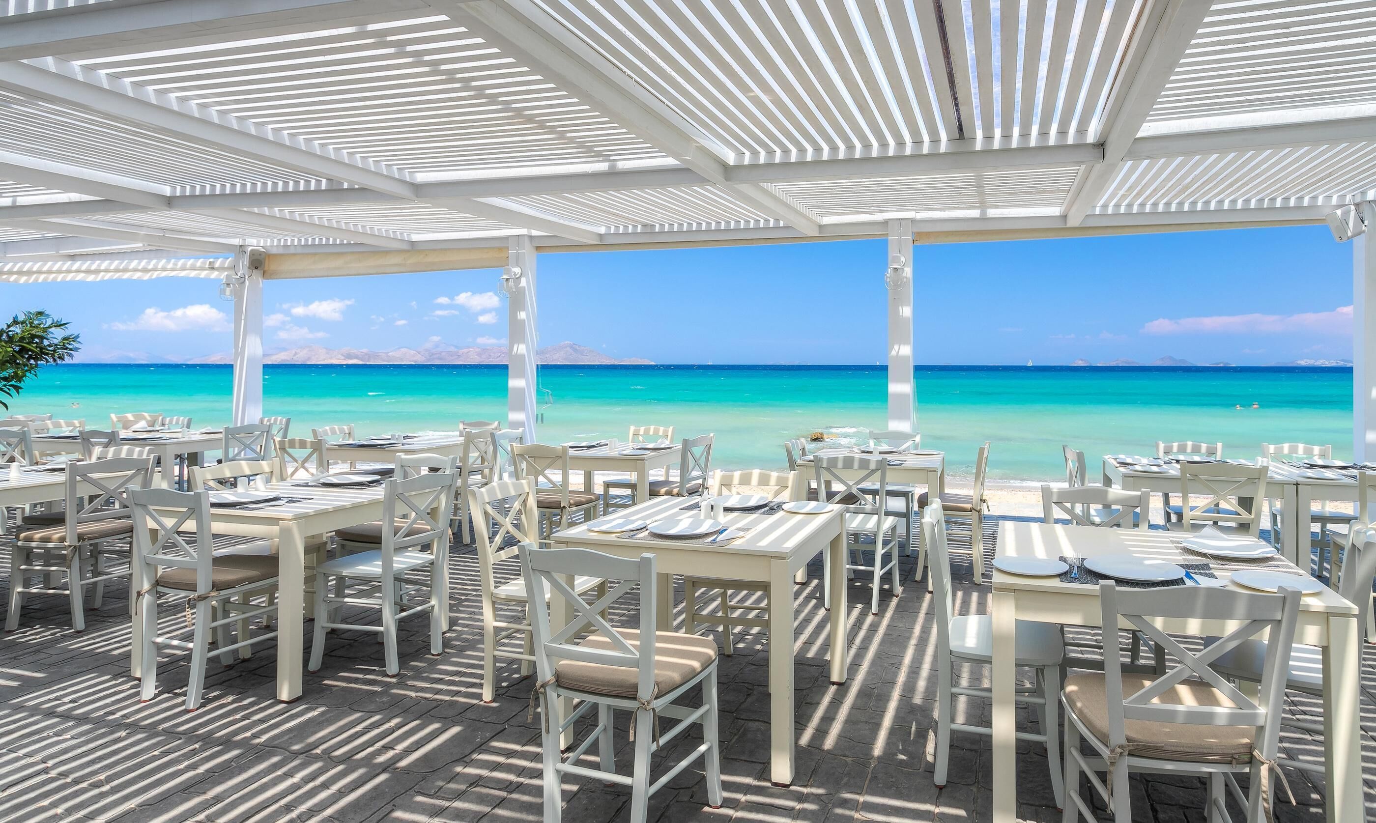 An outdoor dining area with tables and chairs, overlooking a turquoise sea and clear sky.