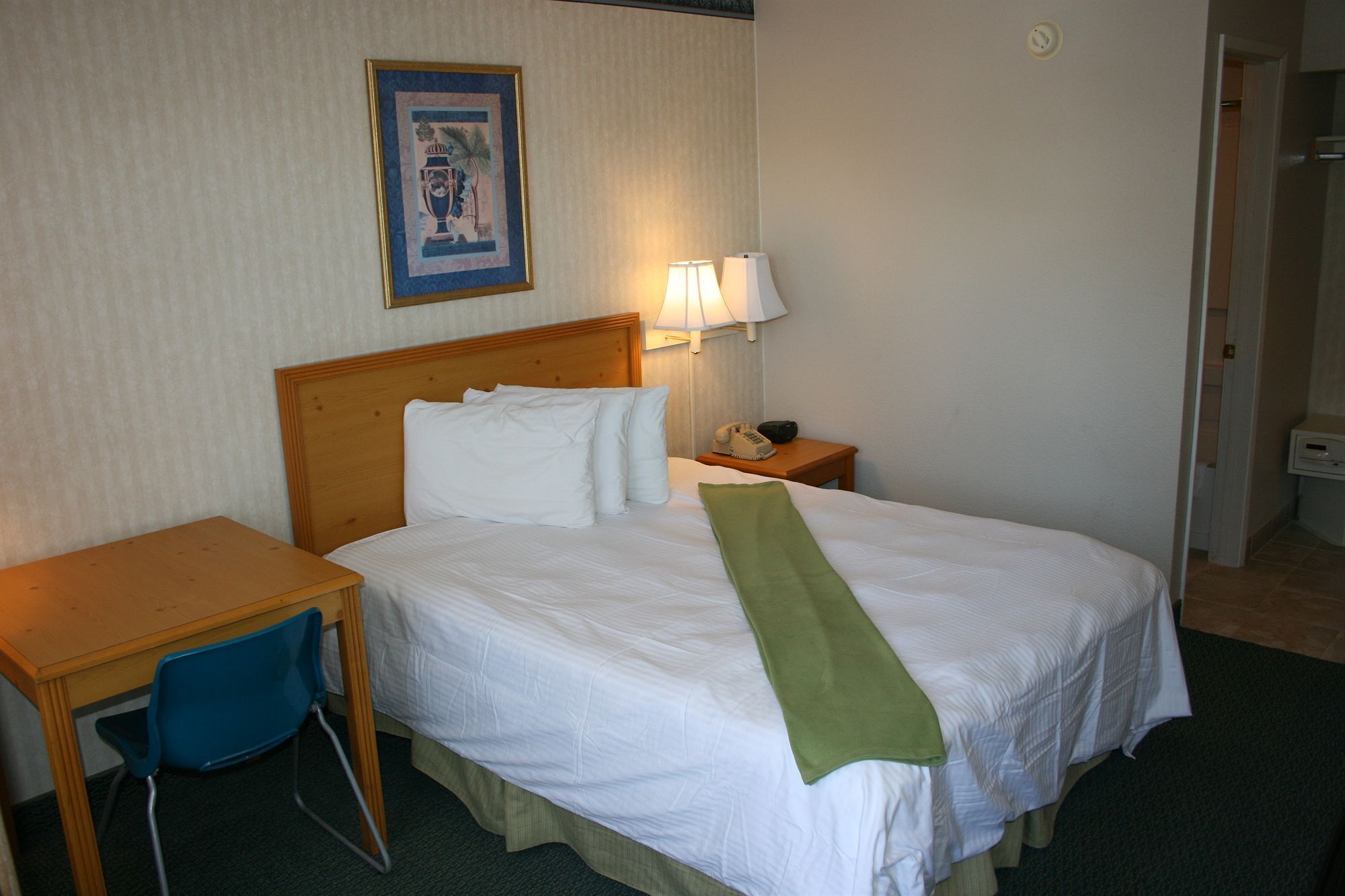 Une chambre d’hôtel avec un lit, un bureau, une chaise, une table de chevet avec une lampe et un tableau encadré au mur.