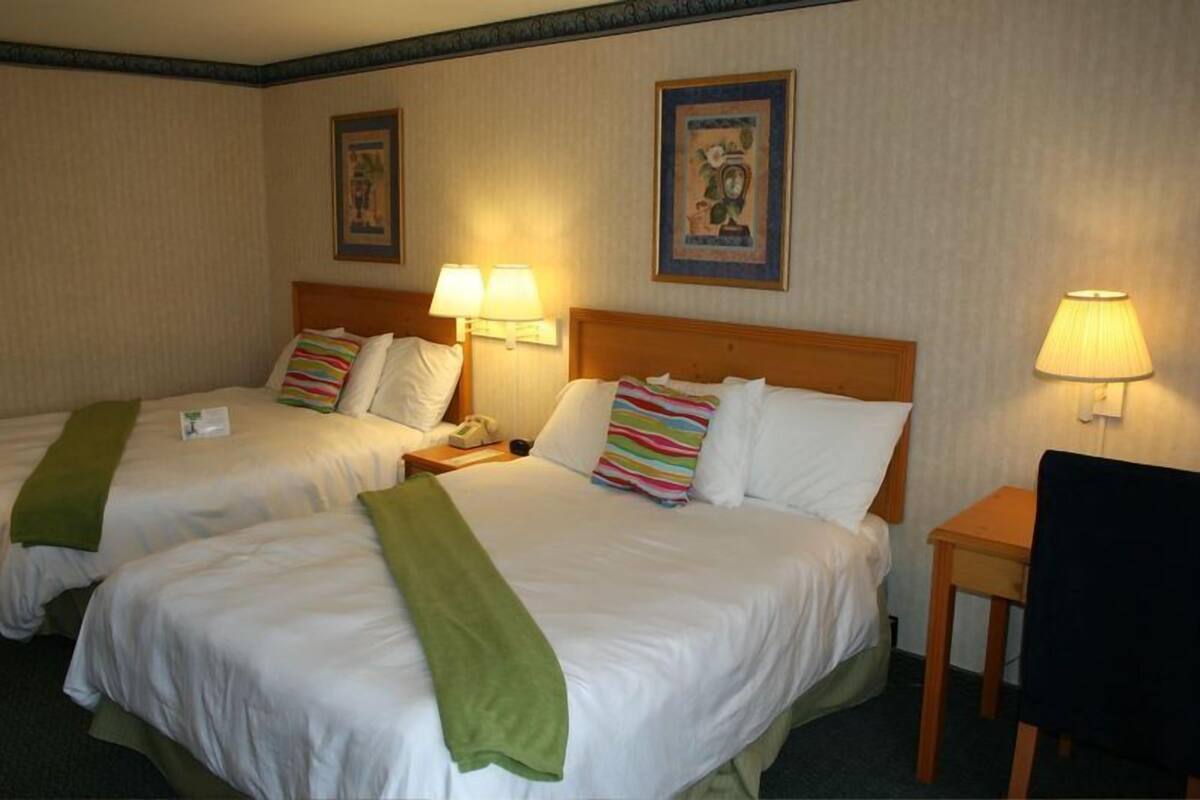 A hotel room with two beds, each with a green throw blanket, bedside lamps, and framed artwork on the walls.