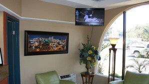 A room with a television mounted on the wall, a framed cityscape picture, a potted plant, and two green chairs. - Bay Inn and Suites at Seaworld (San Diego)