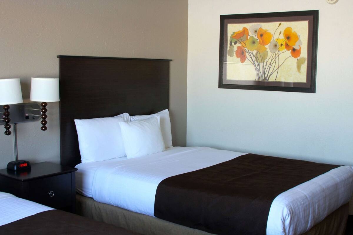 A hotel room with a bed, two bedside lamps, a digital clock, and a framed floral artwork on the wall.