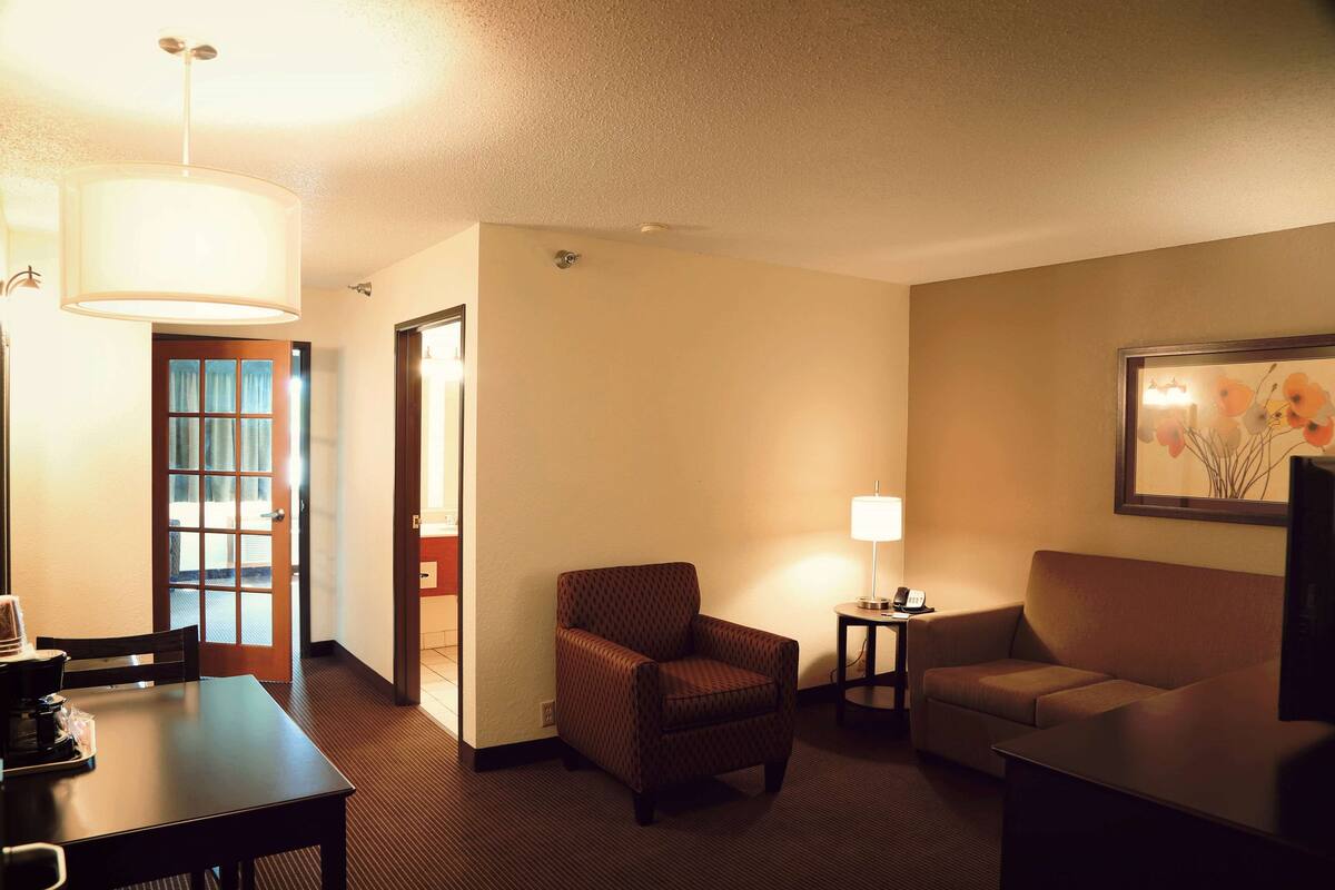 A hotel room with a dining table, two armchairs, a sofa, a lamp, and a painting on the wall.