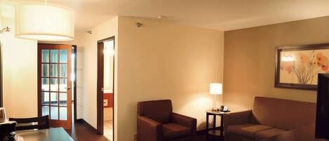 A hotel room with a dining table, two armchairs, a sofa, a lamp, and a painting on the wall.
