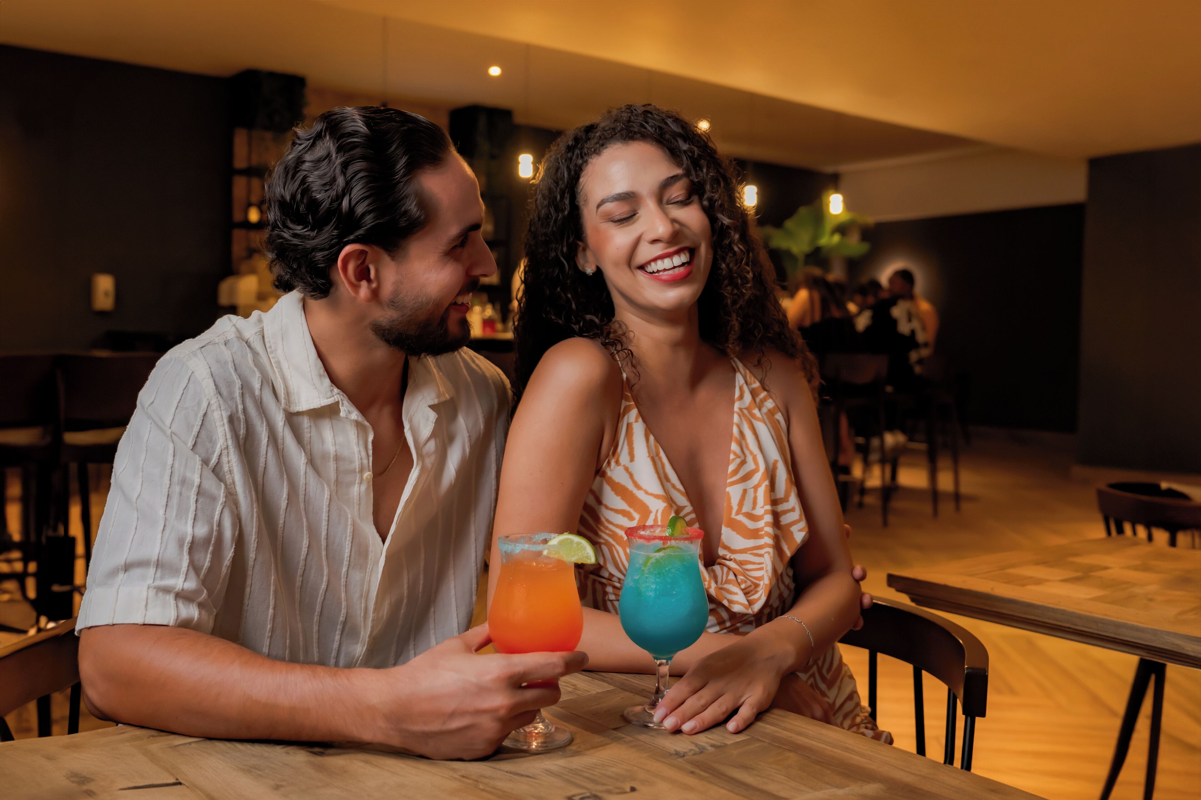 Ein Paar sitzt an einem Holztisch in einer schummrigen Gaststube, möglicherweise ein Restaurant oder eine Bar, mit einer Bar im Hintergrund.
