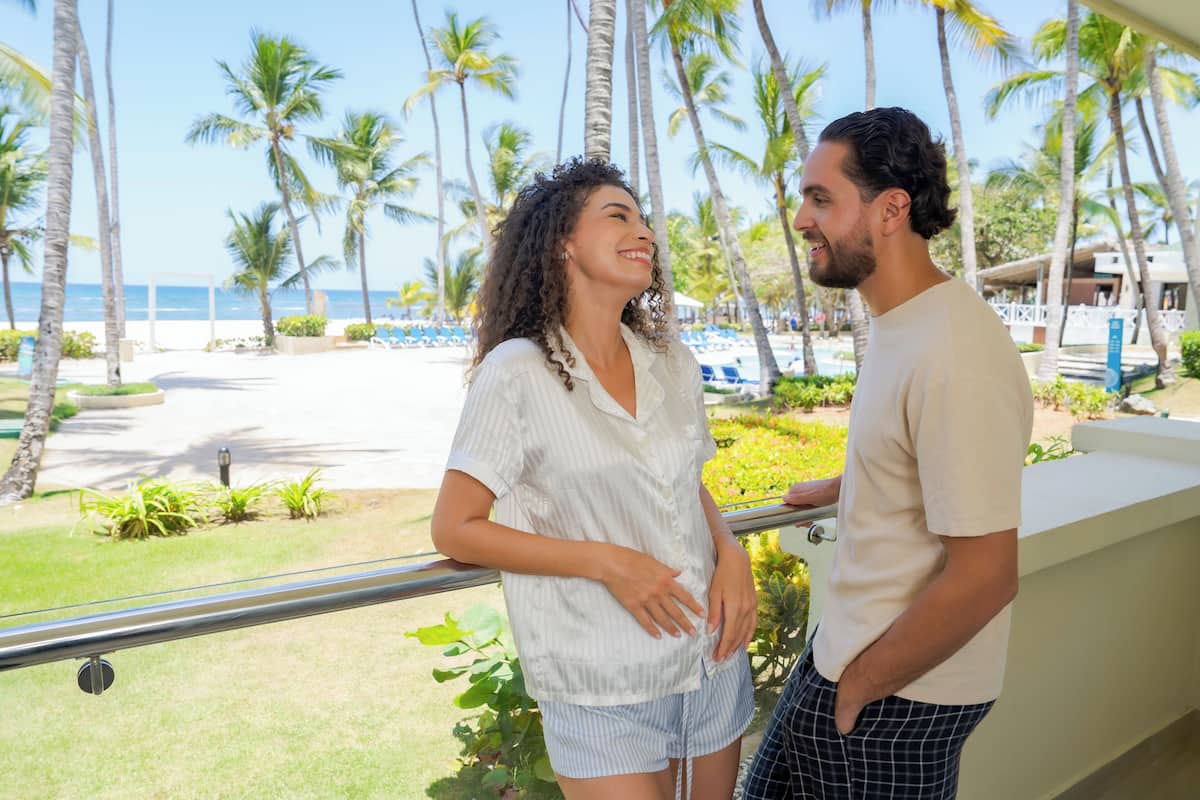 Un couple se tient sur un balcon donnant sur une plage bordée de palmiers et surplombant une piscine.