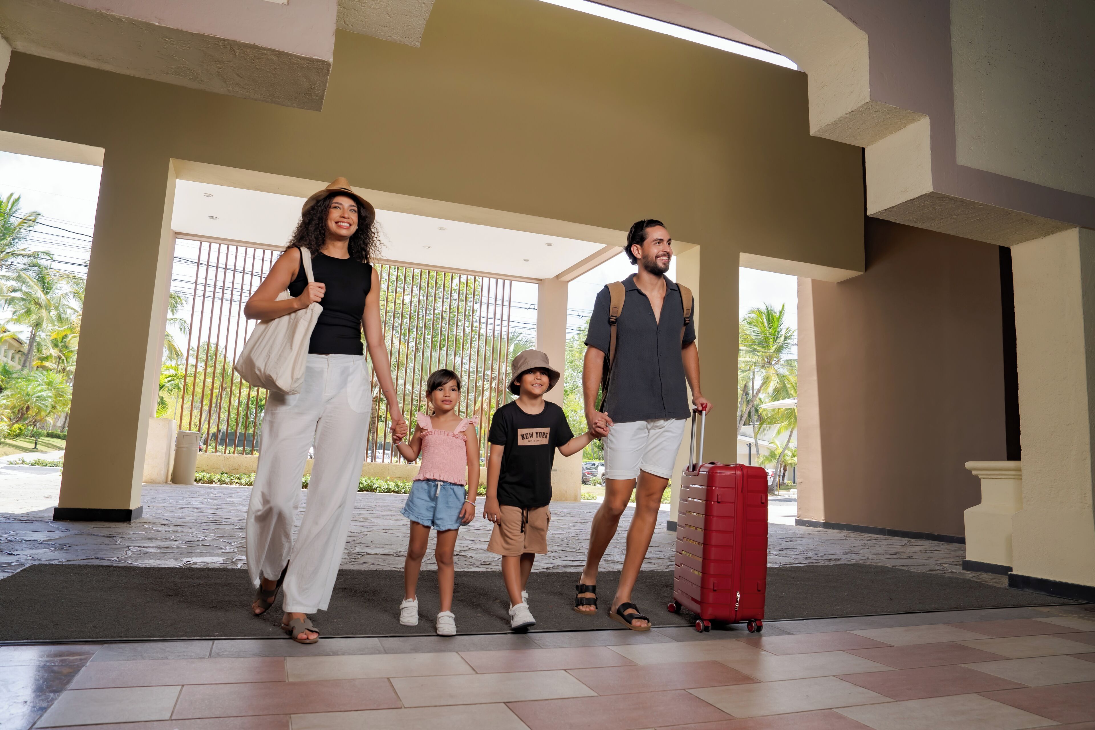 Une famille traverse le hall d’un hôtel, une valise rouge à la main et un enfant portant un haut rose.