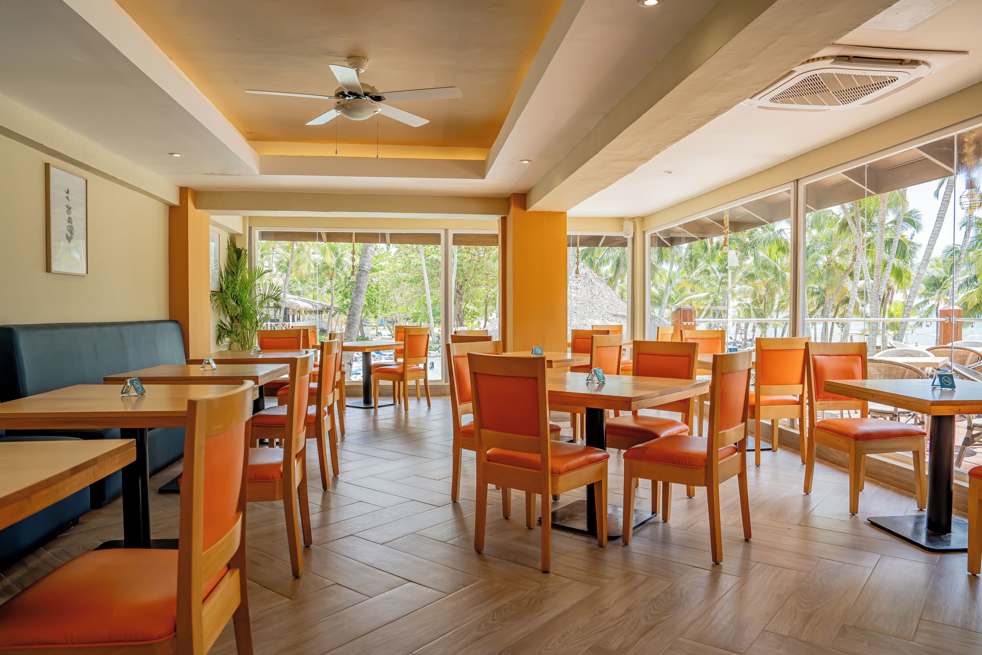 A restaurant with wooden tables and orange chairs, large windows with a view of palm trees, and a ceiling fan.