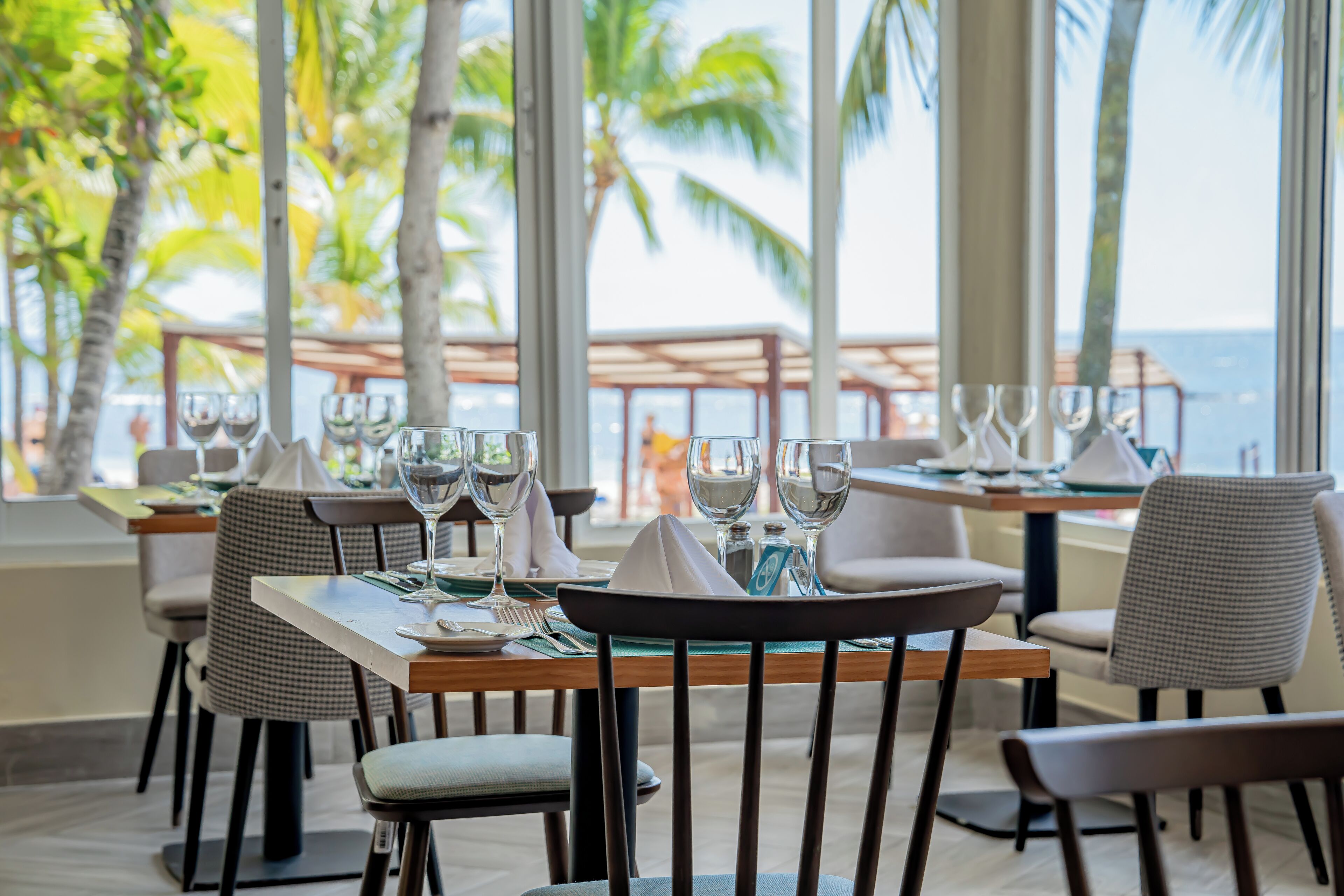 Un restaurante con mesas dispuestas para comer, amplias ventanas que ofrecen vistas a palmeras y al mar.