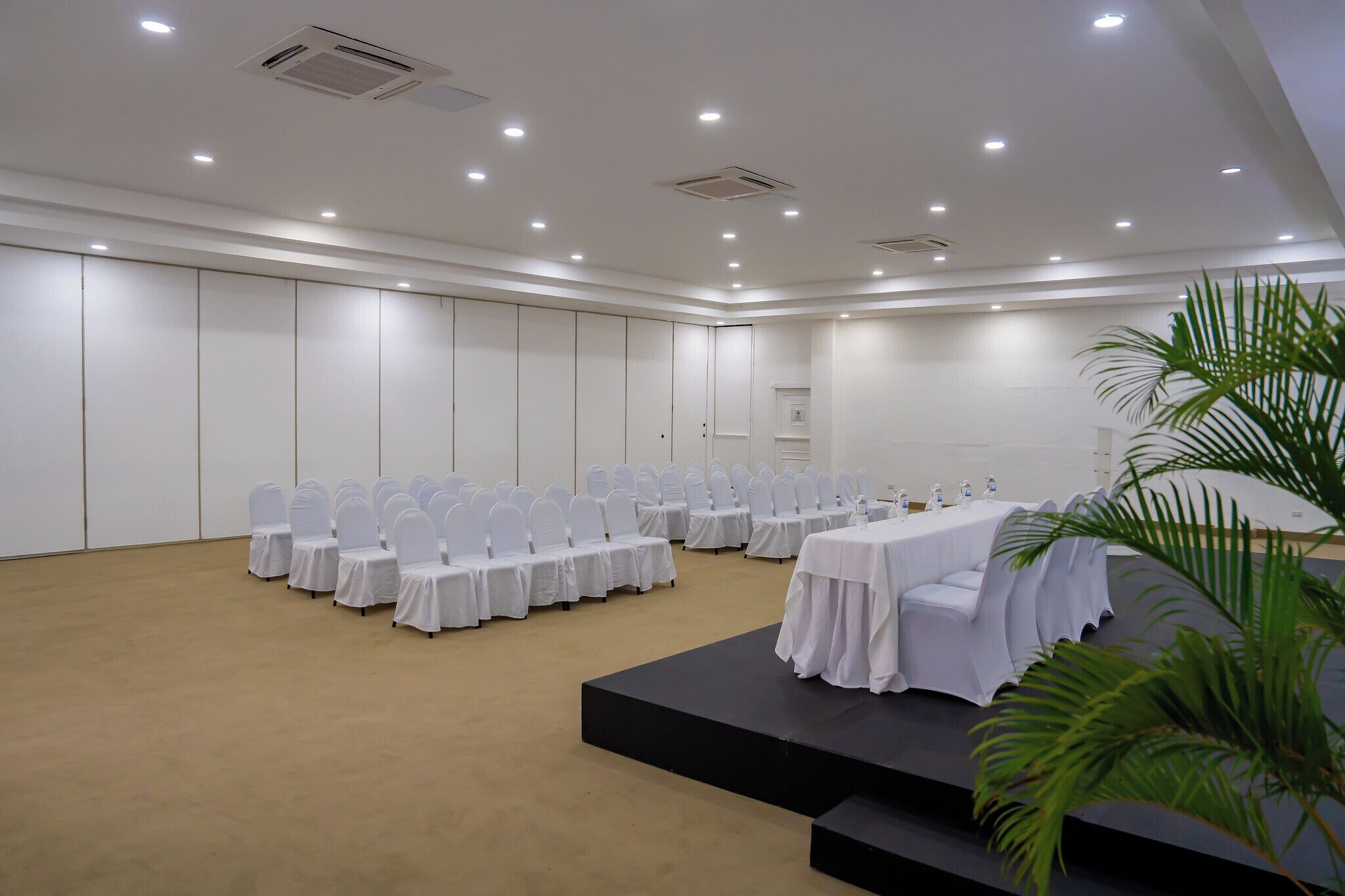 A conference room with rows of white chairs and tables, a stage, and a beige carpet.