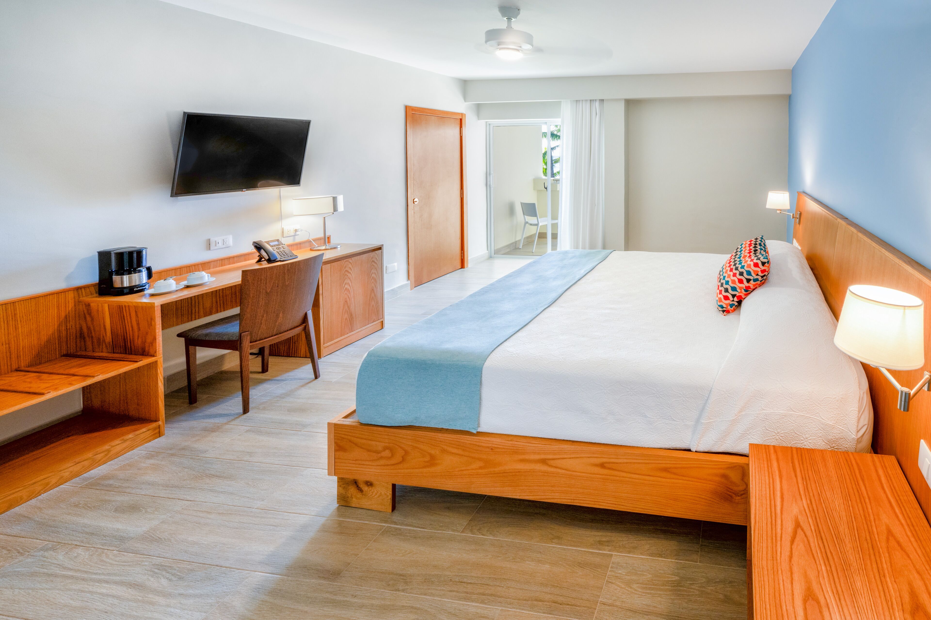 Una habitación de hotel con una cama, un escritorio con silla, un televisor y un ventanal con cortinas.