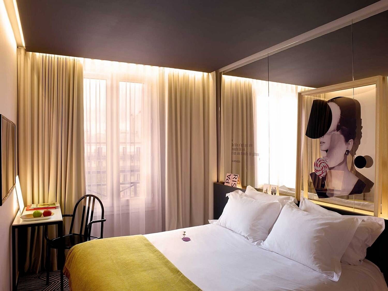 A hotel room with a bed, a desk with a chair, a window with curtains, and a framed picture on the wall.