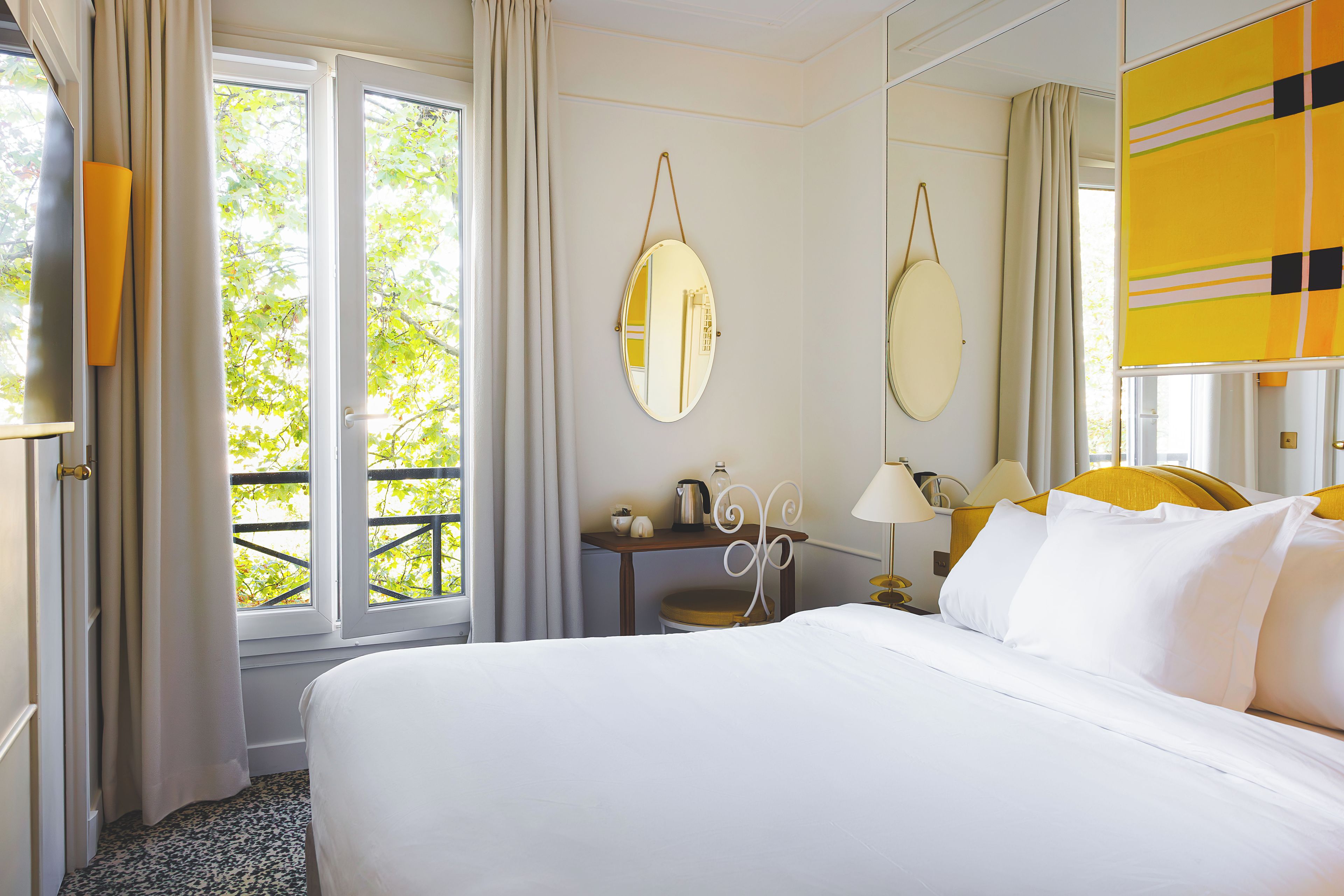A hotel room with a bed, a desk, a chair, a table, a lamp, a mirror, and a window with curtains.