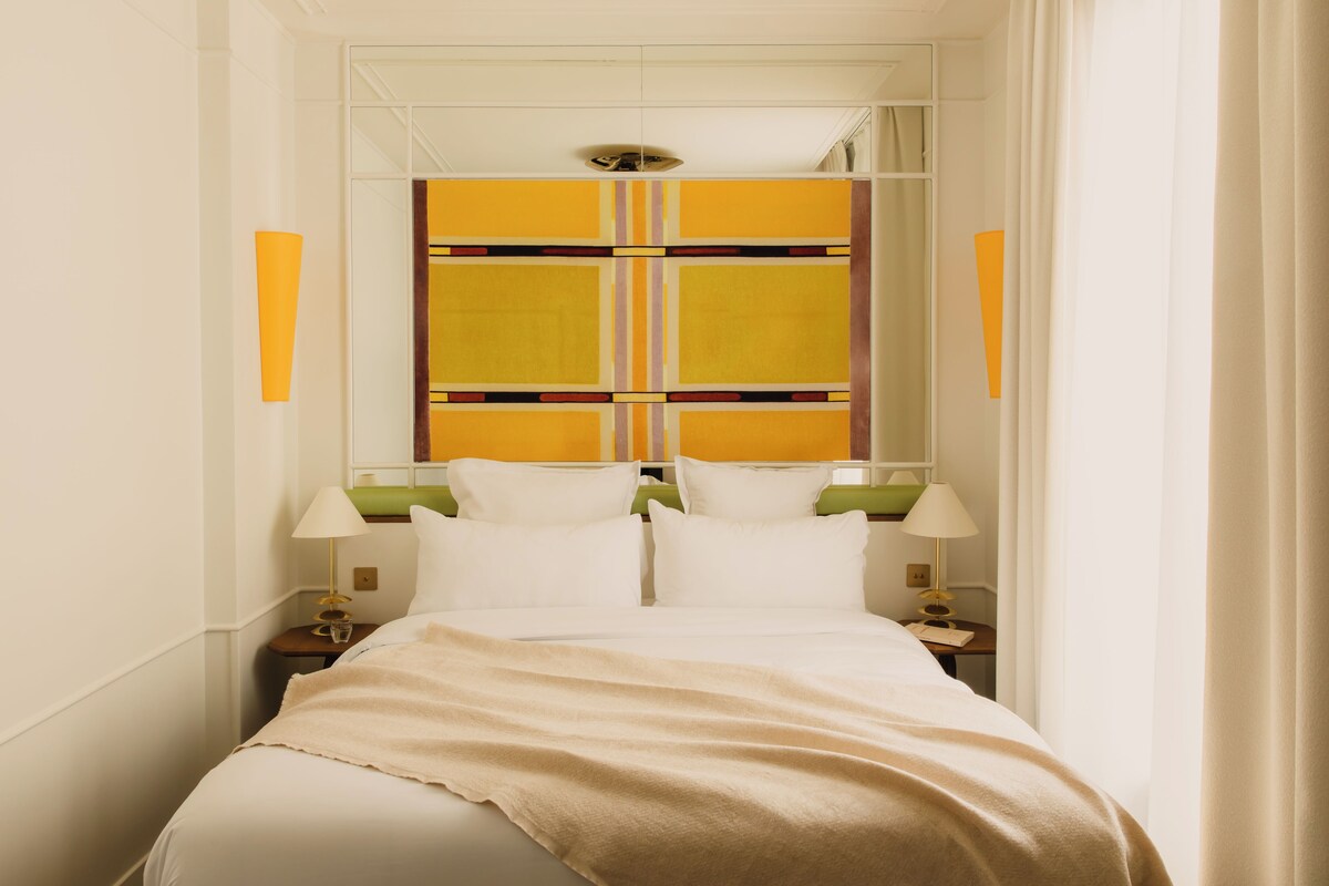A hotel room with a bed, two bedside tables, a nightstand, a window with curtains, and a wall with a decorative panel.