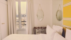 A hotel room with a bed, a window with curtains, a mirror, and a view of buildings. - Hôtel Blanca (Paris)