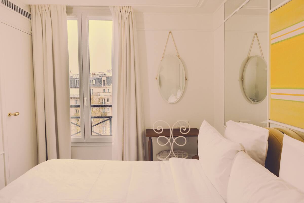 A hotel room with a bed, a window with curtains, a mirror, and a view of buildings.