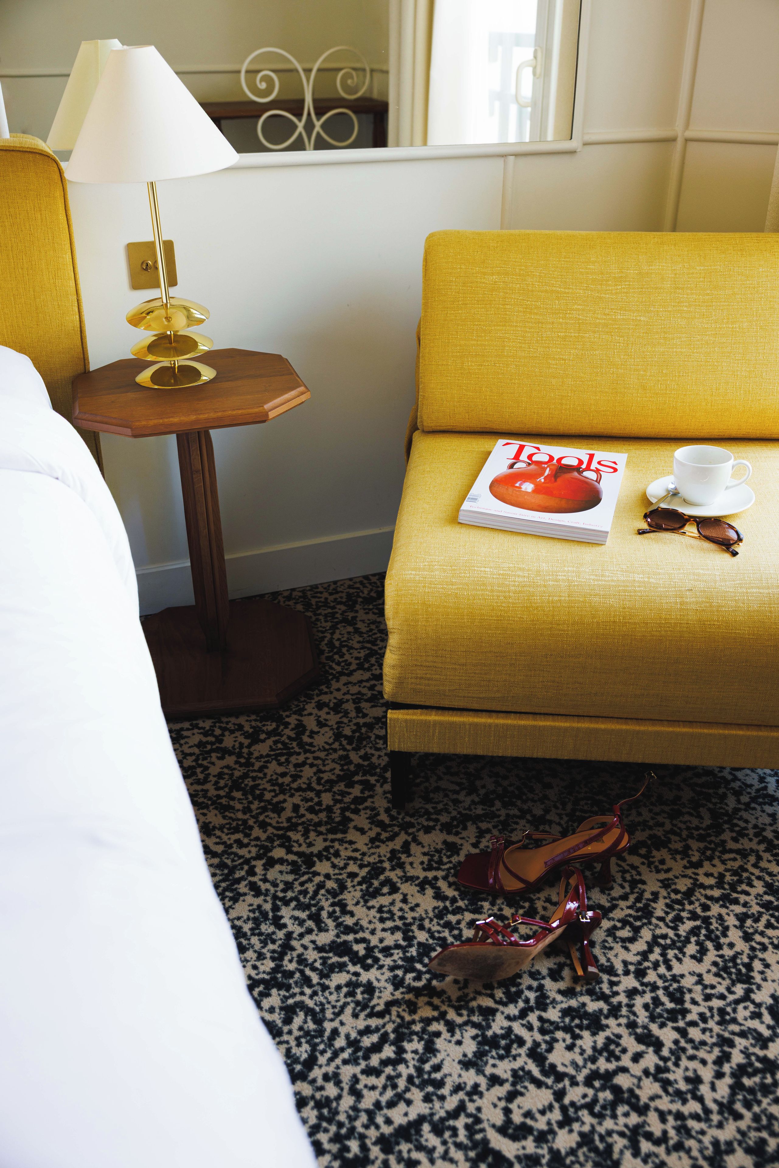 Habitación de hotel con una cama, una mesita de madera con una lámpara, un sillón amarillo con una revista, una taza blanca y un par de gafas de sol.