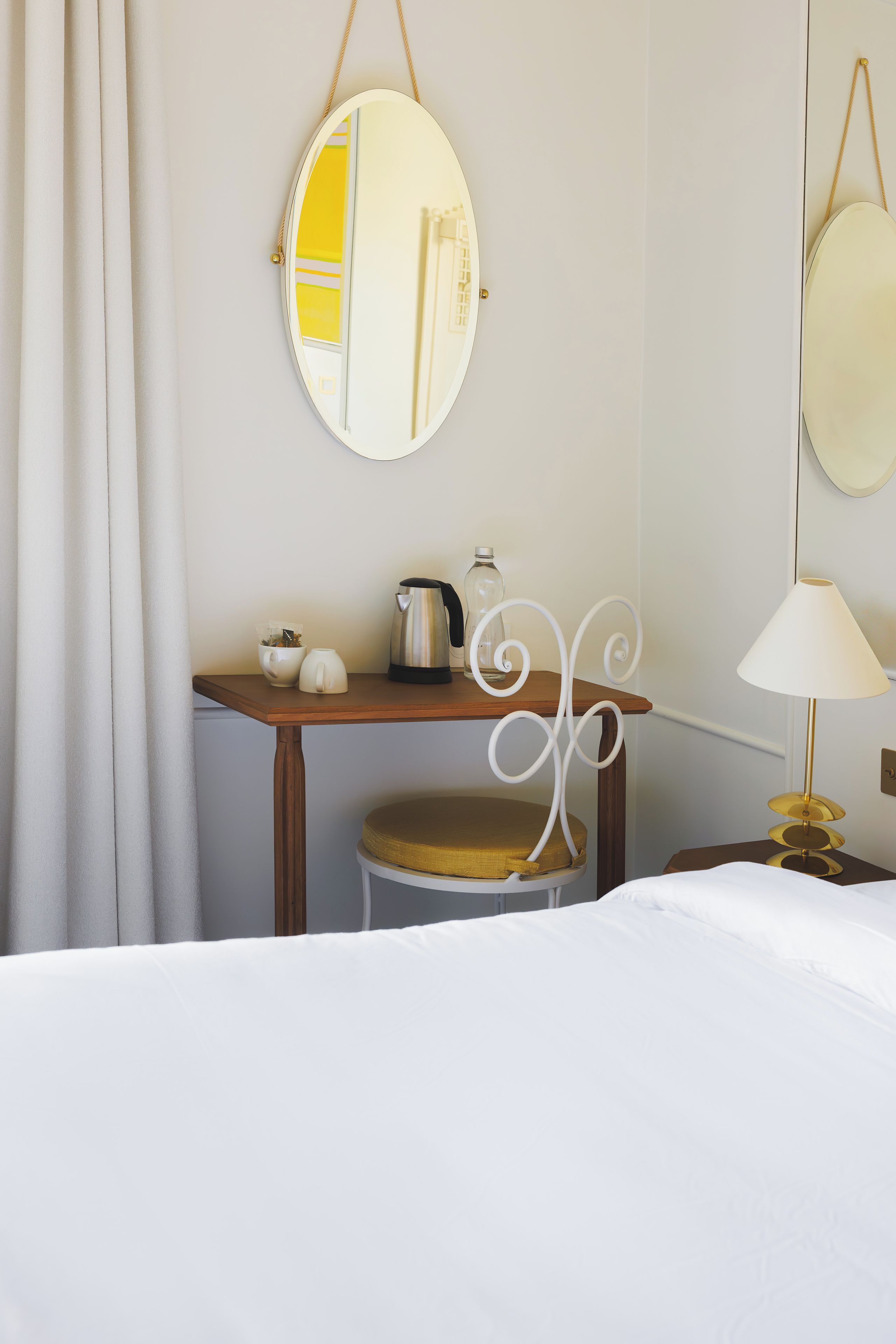A hotel room with a bed, a small table with a kettle, a cup, and a bottle, and a chair with a cushion.