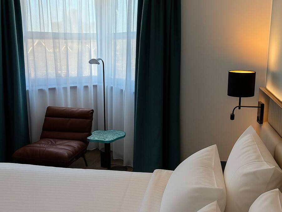 Habitación de hotel con una cama grande, un sillón de cuero marrón, una mesita, una lámpara y vistas al exterior a través de cortinas traslúcidas.