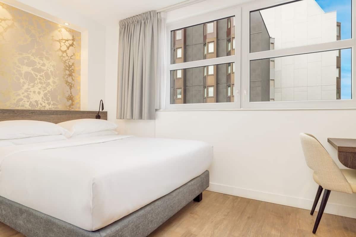 Une chambre d’hôtel comprenant un lit, un bureau, une chaise, une fenêtre donnant sur des immeubles et un mur orné d’un motif texturé.