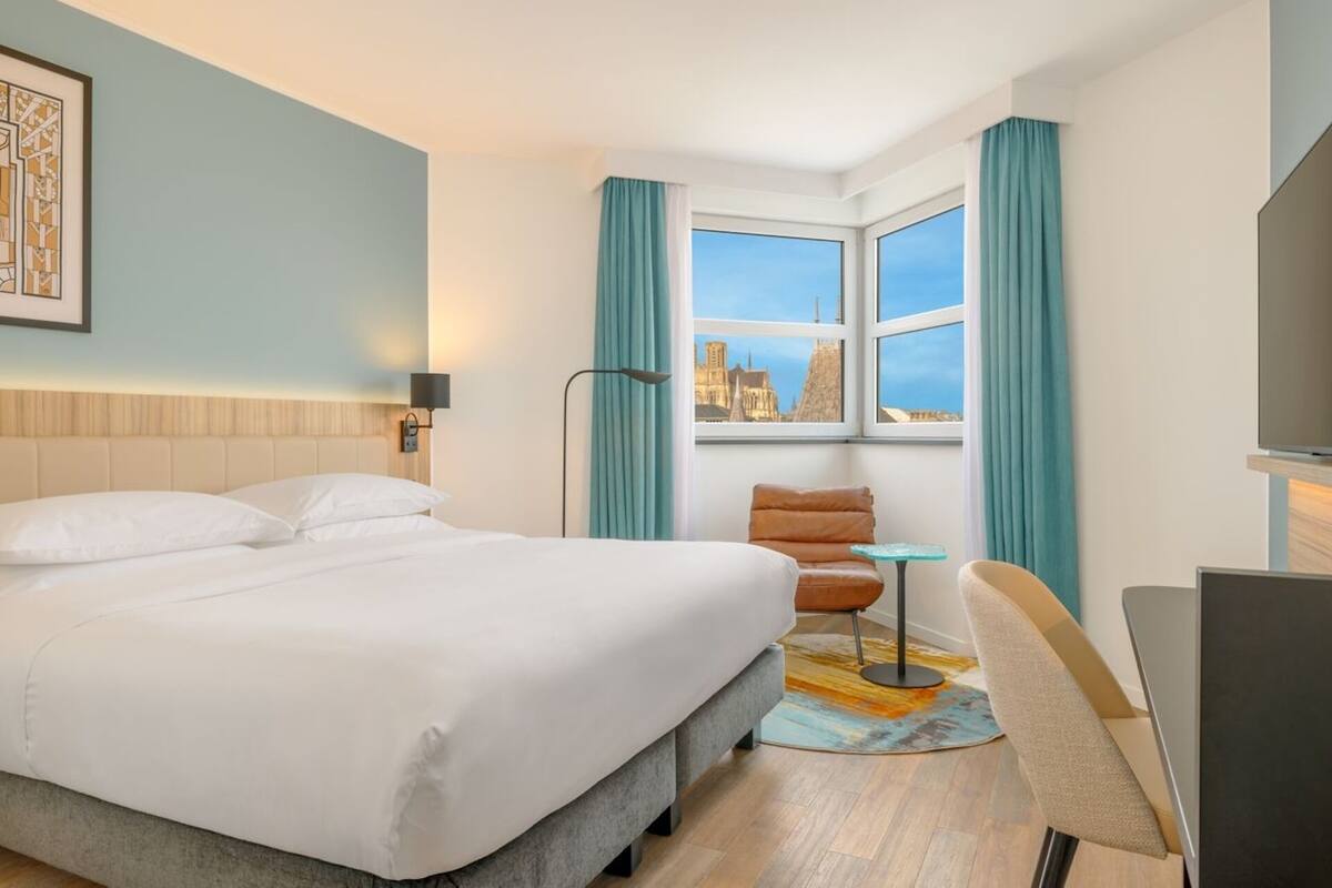 Une chambre d’hôtel comprenant un lit, un bureau, une chaise, une télévision, une fenêtre avec des rideaux et une vue sur la ville.