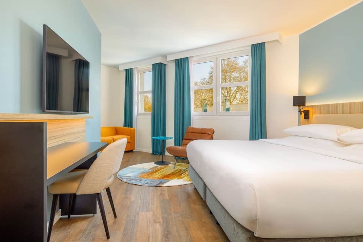 Une chambre d’hôtel avec un lit, un bureau, une chaise, une télévision et une fenêtre avec des rideaux.