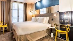 Una habitació d'hotel amb un llit gran, una taula de vidre, una cadira groga i una vista de la ciutat per la finestra.