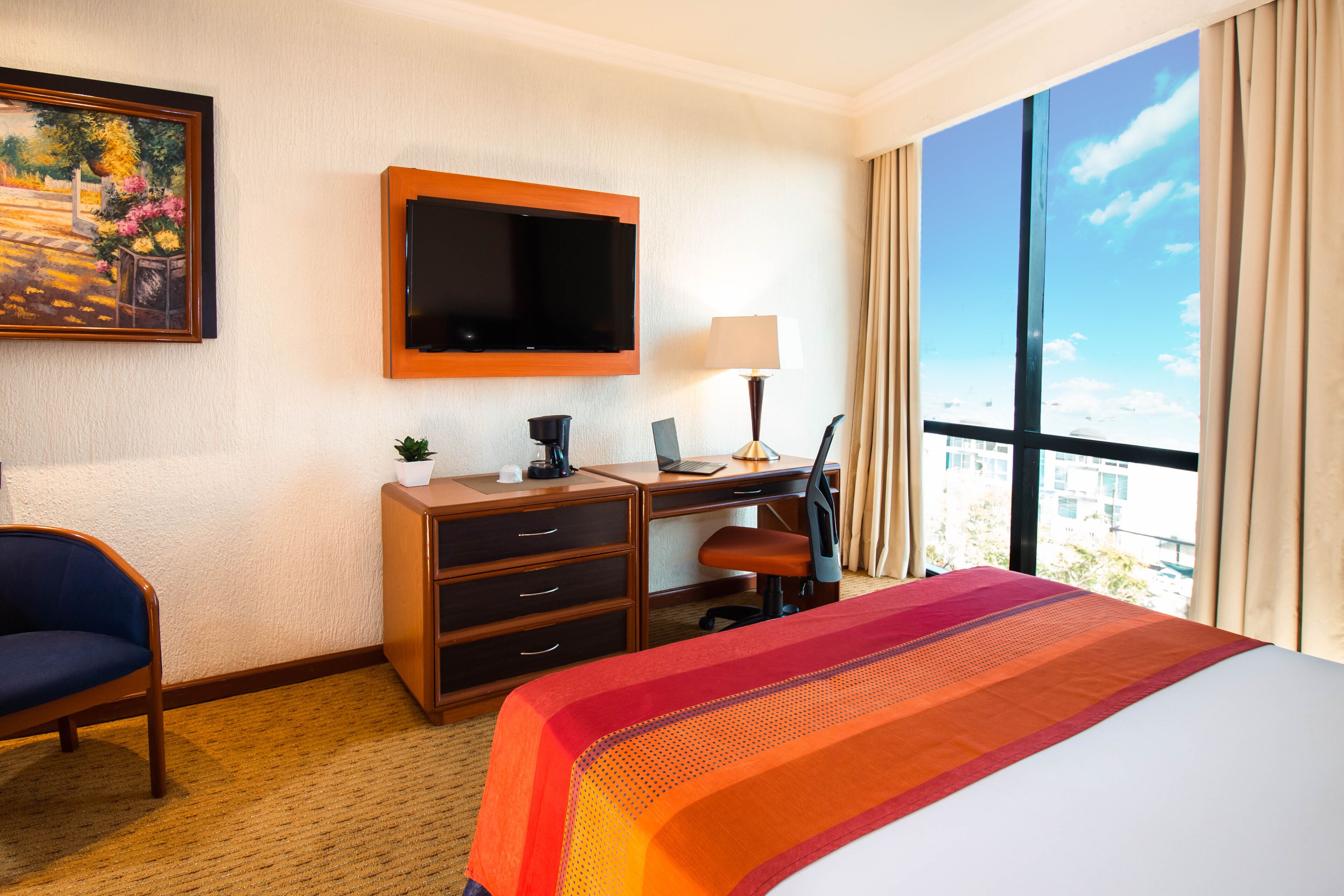 Habitación de hotel con cama, escritorio con computadora, silla, televisor montado en la pared y ventana con cortinas.