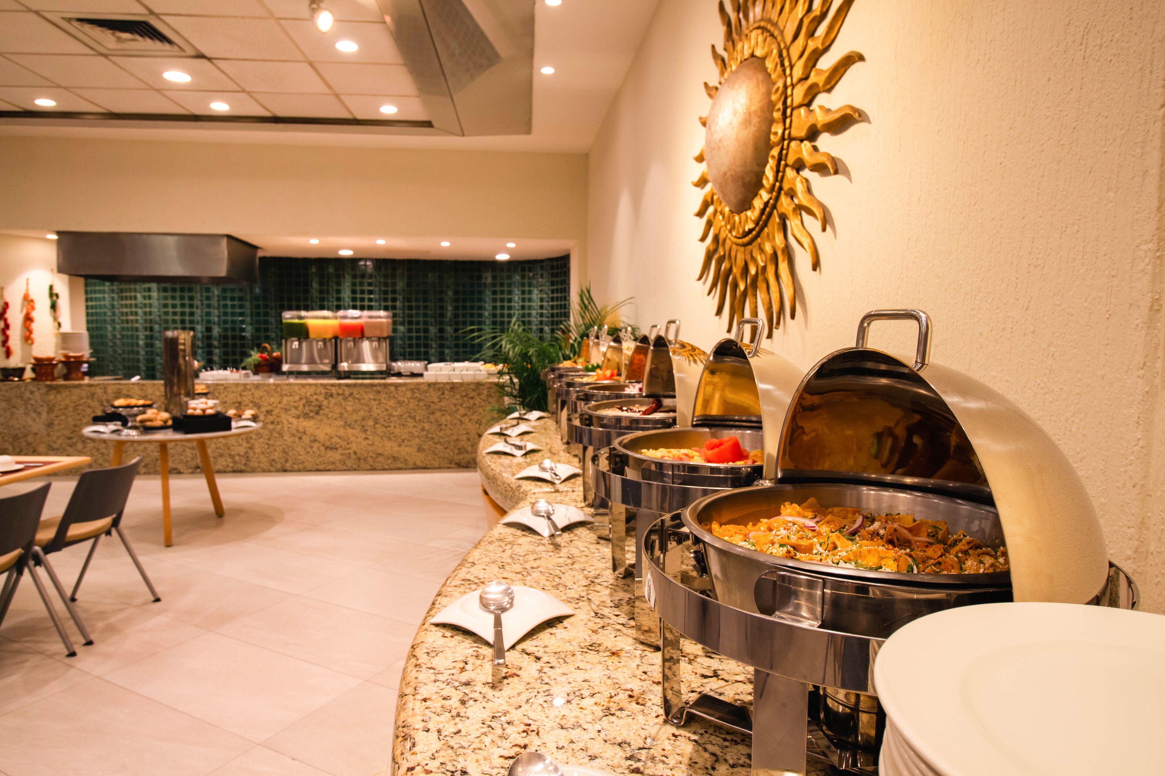 A buffet setup with various food stations, a counter with a coffee machine, and a decorative sun-like wall piece.