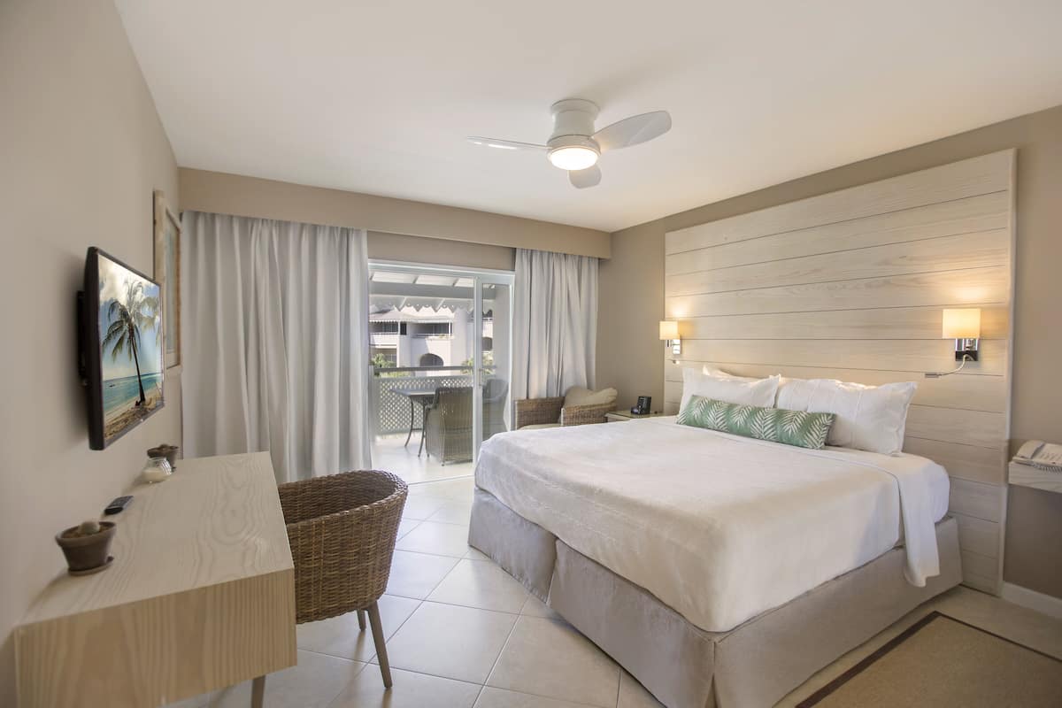 A hotel room with a bed, a desk, a chair, a ceiling fan, and a view of the outside.