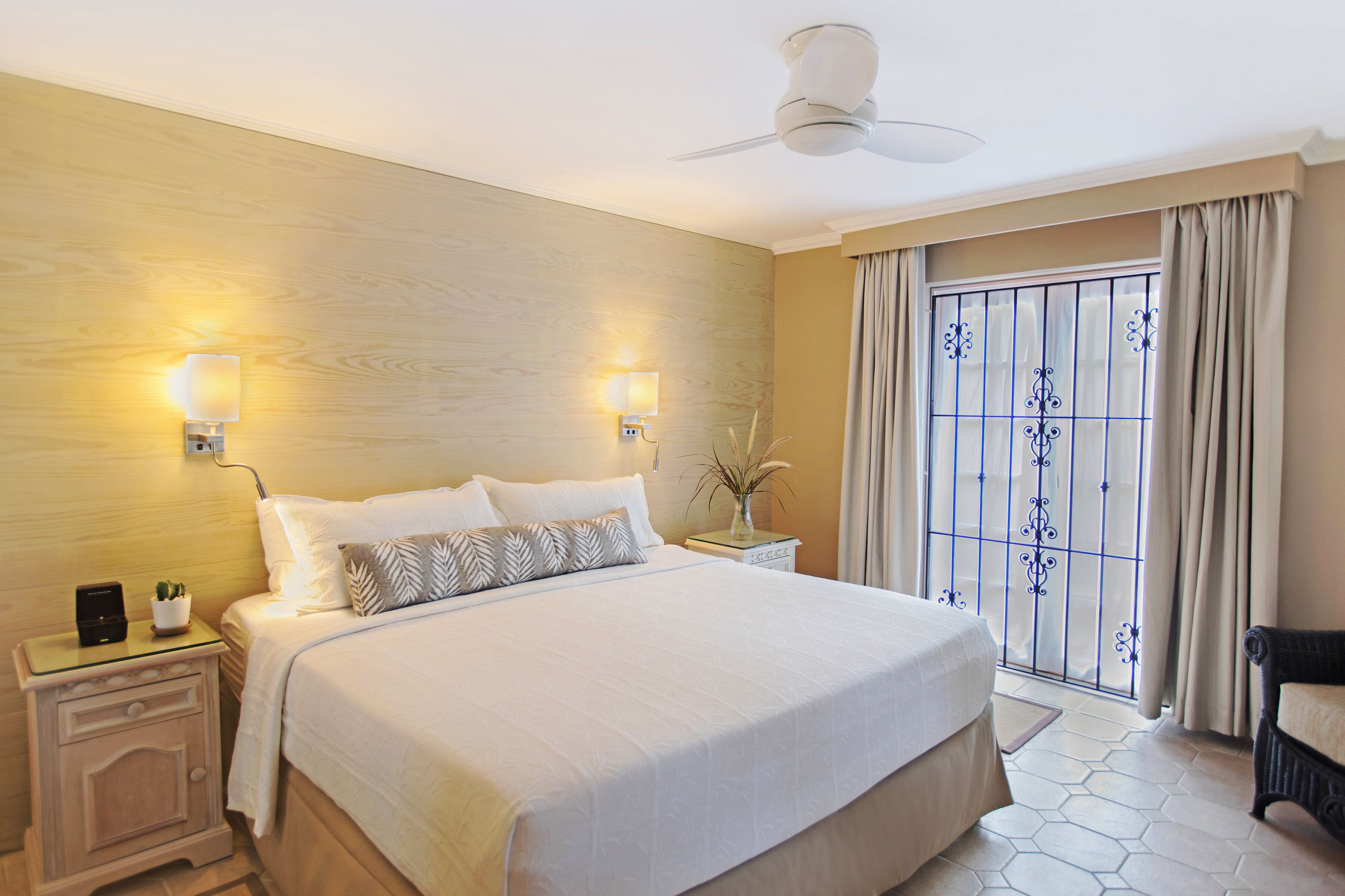 Een hotelkamer met een groot bed, twee bedlampen, een nachtkastje, een plafondventilator en een raam met decoratieve gordijnen.