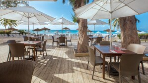 An outdoor dining area with wooden tables and chairs, white umbrellas, and a view of the beach and ocean. - Bougainvillea Barbados (Maxwell)