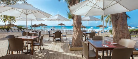 Un espace de restauration en plein air avec des tables et des chaises en bois, des parasols blancs et une vue sur la plage et l’océan.