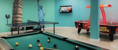 Een pooltafel met ballen, een tafeltennistafel en een televisiescherm in een kamer met turquoise muren en een plafond met een Union Jack ontwerp.