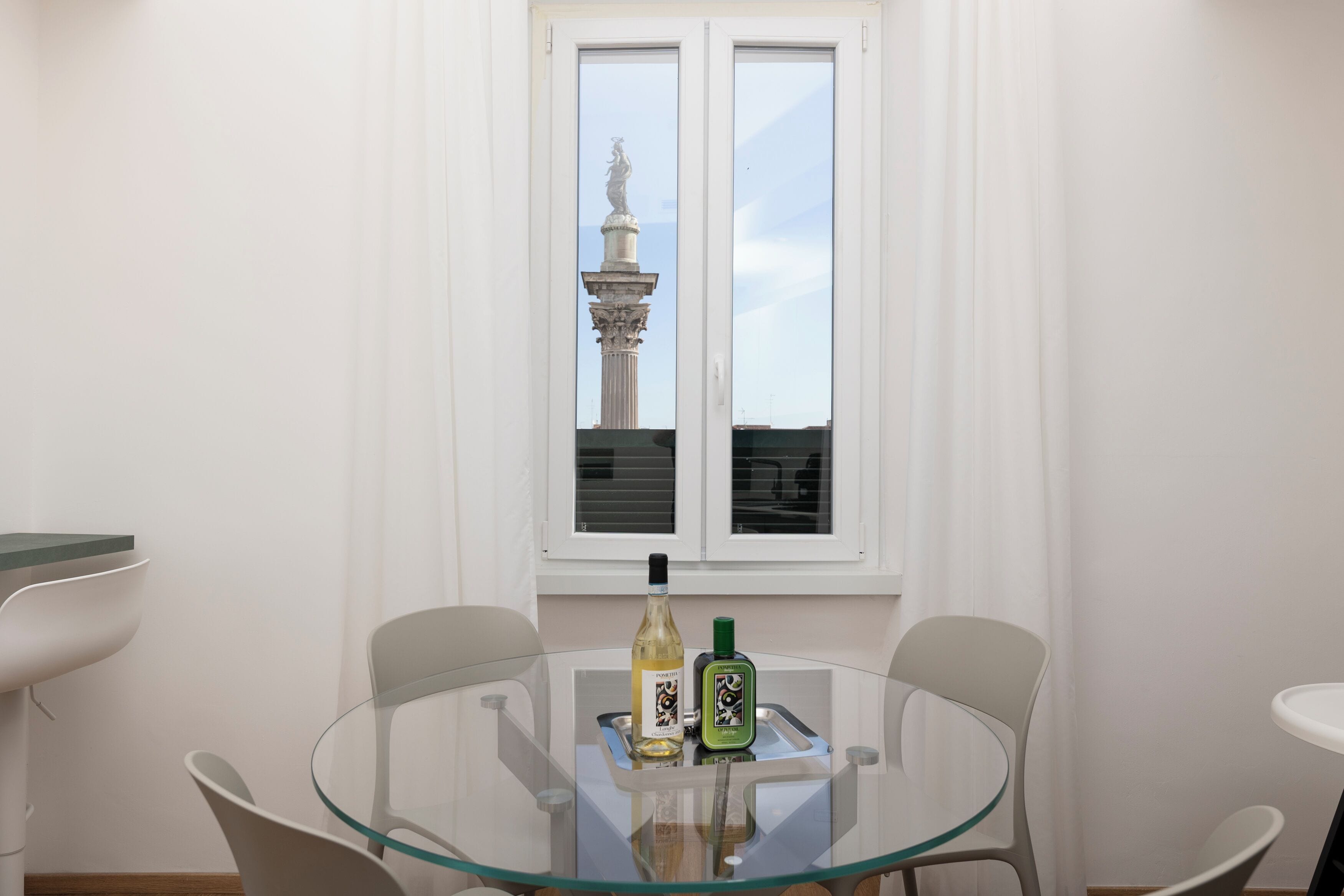Une pièce avec une table ronde en verre, deux chaises, une fenêtre donnant sur une statue et deux bouteilles posées sur la table.