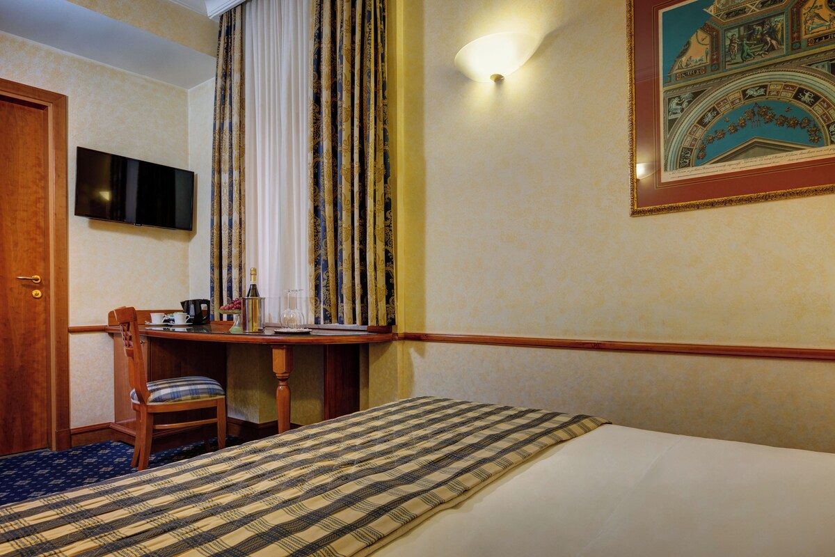 A hotel room with a bed, a desk with a chair, a television, and a framed picture on the wall.