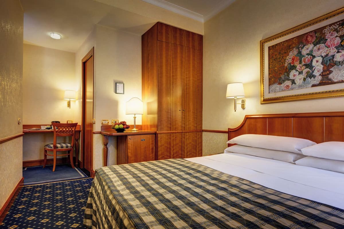 A hotel room with a bed, a desk, a chair, a wardrobe, and a painting on the wall.