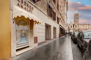 A hotel facade with a prominent entrance, a clock tower in the background, and parked cars along the street.