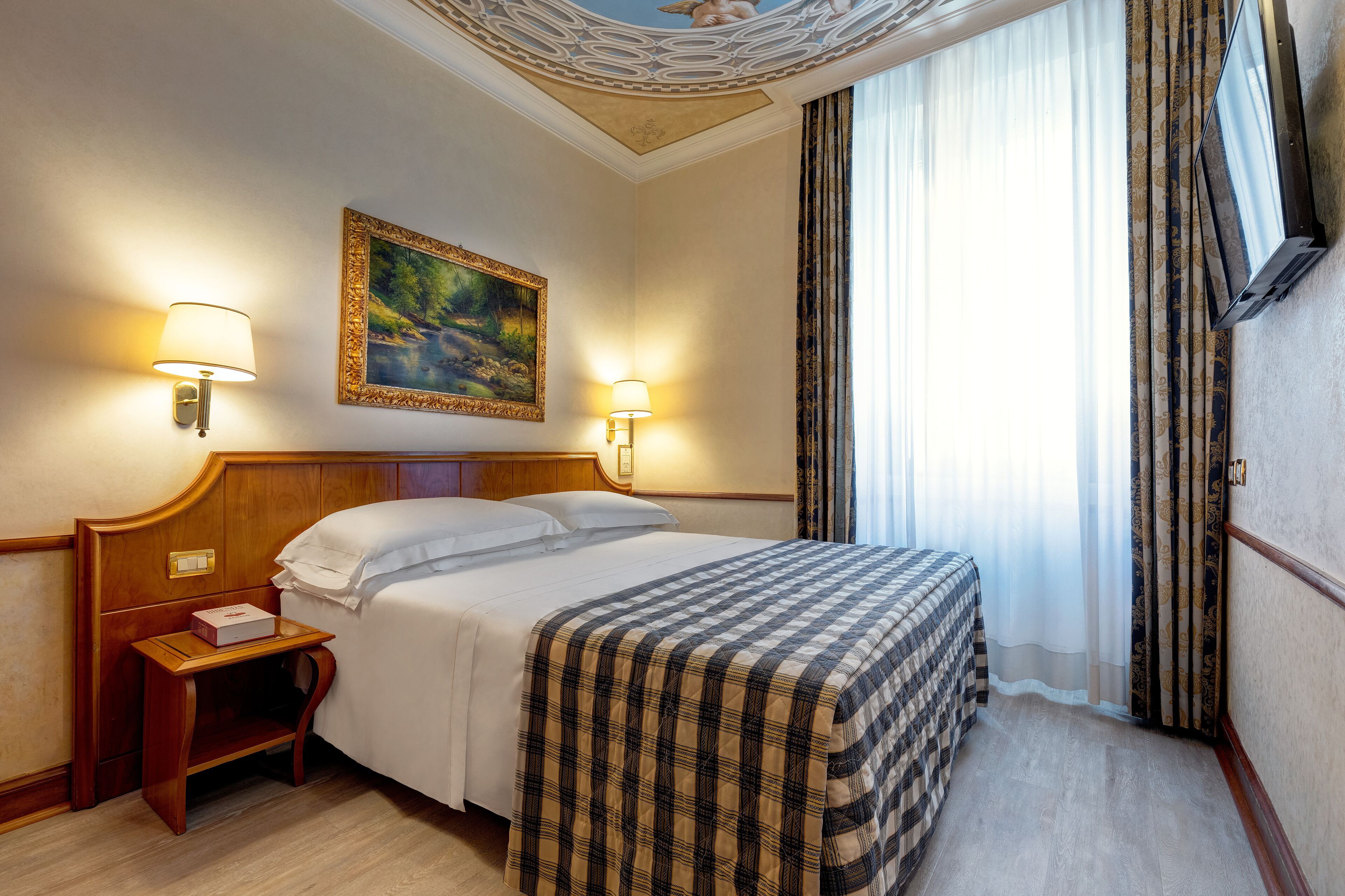 Habitación de hotel con cama, dos lámparas de noche, un cuadro en la pared y una ventana con cortinas.