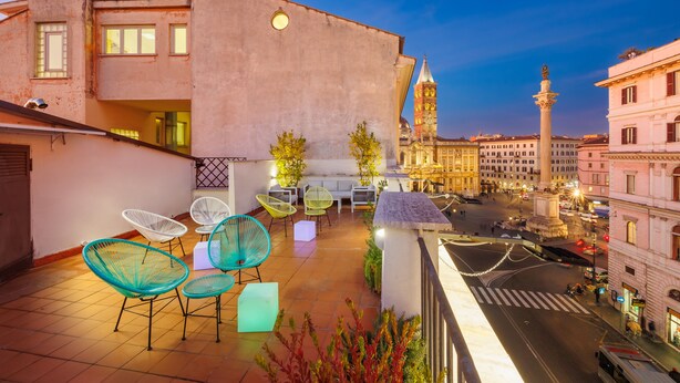 Une terrasse sur le toit avec des sièges en plein air, une vue sur une place historique et un beffroi en arrière-plan.
