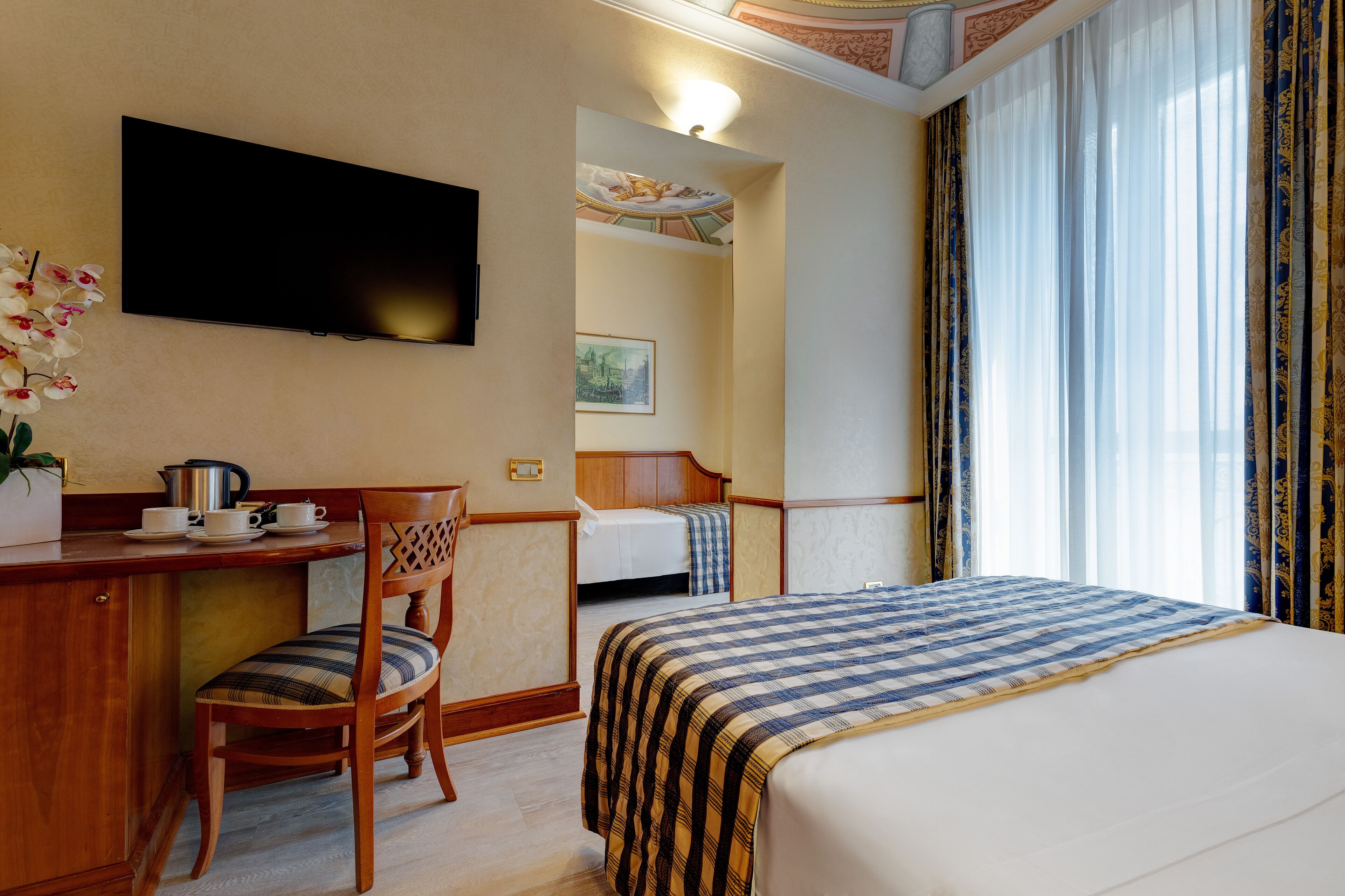 Une chambre d’hôtel avec un lit, un bureau et une chaise, un téléviseur, un tableau au mur et une fenêtre avec des rideaux.