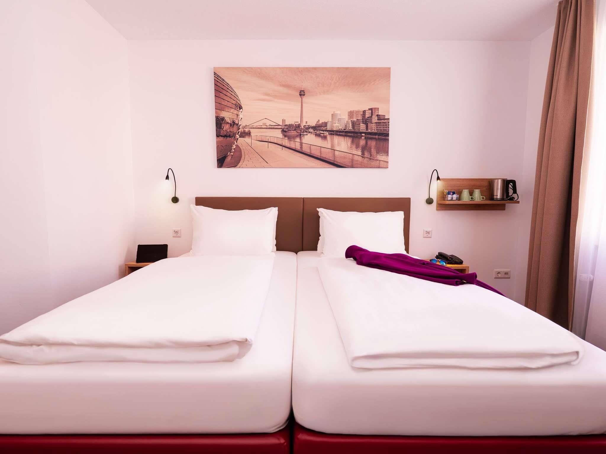 A hotel room with two beds, a cityscape painting on the wall, a bedside table with a lamp, and a shelf with cups and a phone.