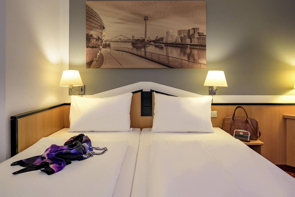 A hotel room with a bed, two wall-mounted lamps, a cityscape painting, and a handbag on the nightstand.
