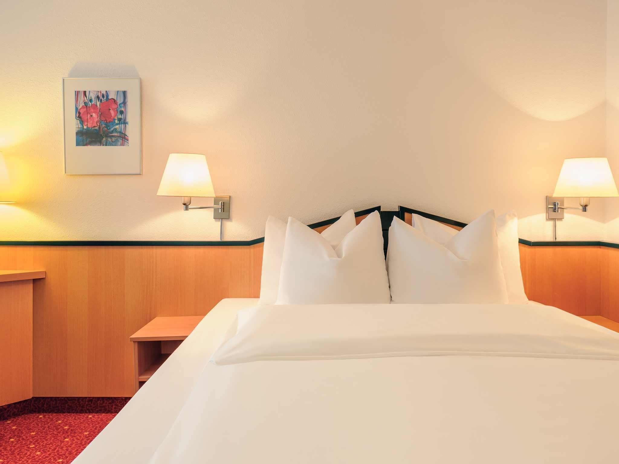 Quarto de hotel com uma cama bem arrumada, duas luminárias laterais, uma mesinha de cabeceira e uma obra de arte emoldurada na parede.